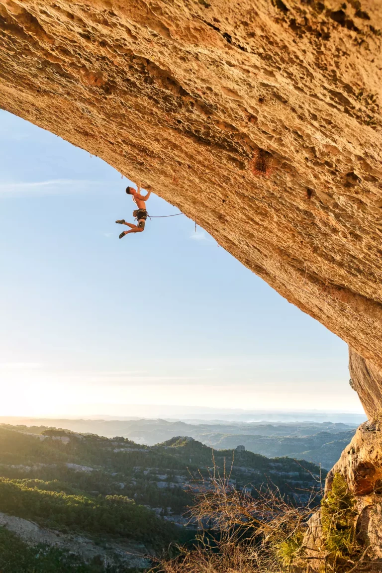 A climber on Elleanor, a steep orange wall of conglomerate rock, with a mountainous landscape in the background at sunset in Margalef.